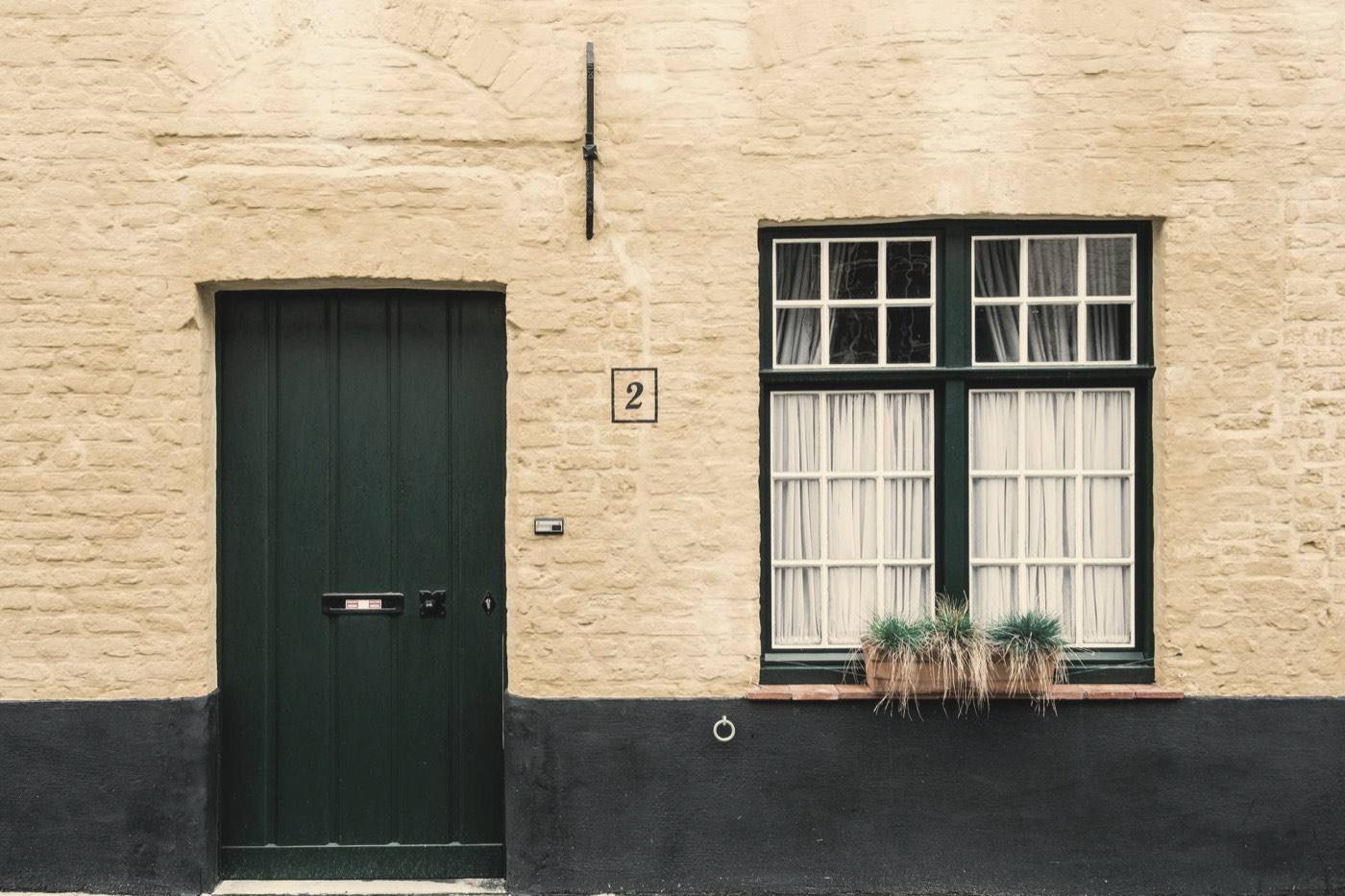 A dark green front door, closed, in a cream painted brick wall beside a curtained sash window with a planter on the sill