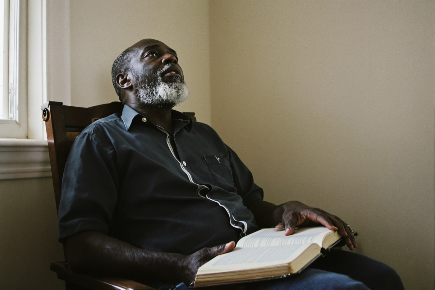 A man sitting in a wooden chair beside a window with a large open book on his lap, looking up in thought