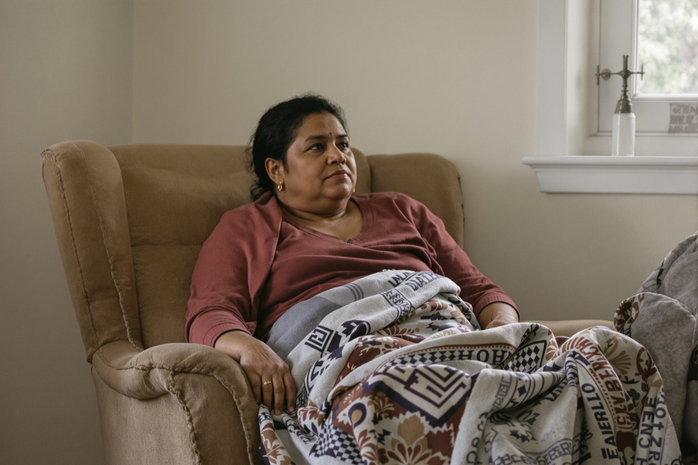 A woman sitting back in an armchair with a patterned blanket across her lap, hands still, looking steadily across the room